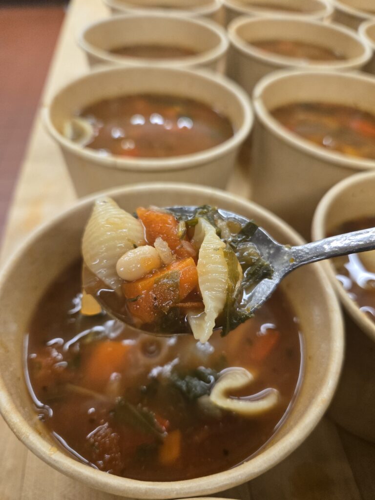A spoonful of vegetable and pasta soup is lifted from a bowl, with several bowls of soup in the background.