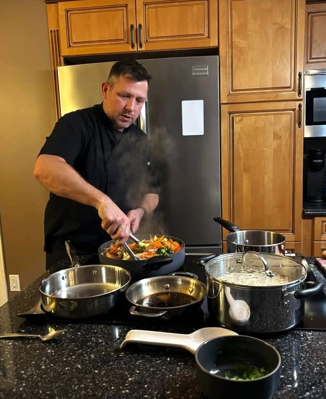 A man is cooking in a kitchen, stirring vegetables in a pan with steam rising, surrounded by various cooking pots.