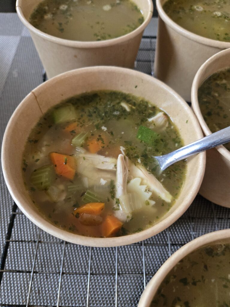 A bowl of soup with chicken, vegetables, and herbs, served in a disposable cup.