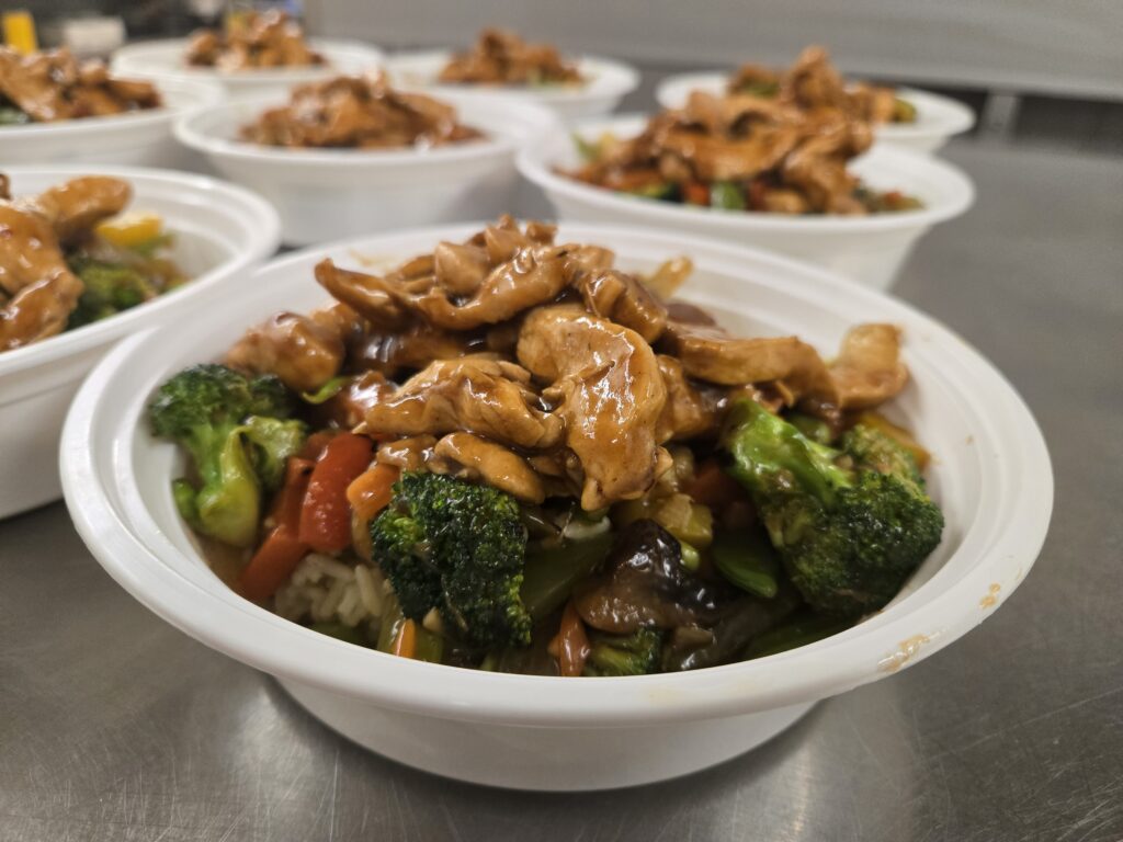 A close-up of a bowl of chicken stir-fry with vegetables over rice, surrounded by similar bowls in the background.